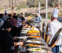 Guests line up at an outdoor buffet of chafing dishes, a chef in a tall white hat overseeing the service.