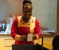 Smiling woman in a red traditional dress hands a welcome card to a guest at a desk with a laptop in a warmly lit reception area.
