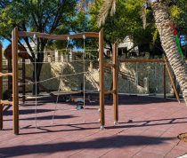 Outdoor playground with wooden climbing frame and rope netting amid palm trees on a brick surface